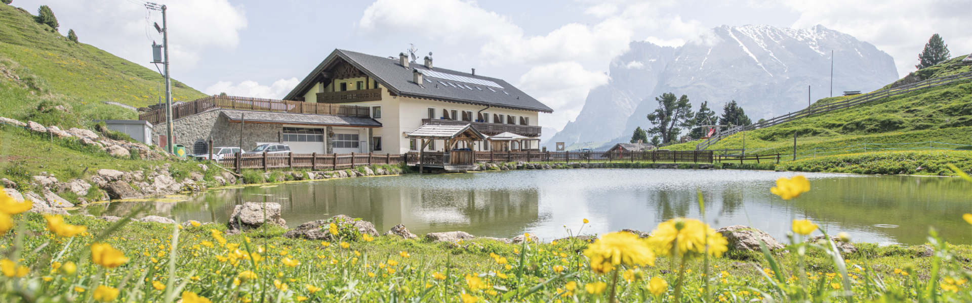 Molignon Alpe di Siusi refuge | The Alpine paradise with flowering meadows and the imposing Sassolungo and Sassopiatto mountains The picture shows the Molignon hut with the lake in the foreground and the Landkofel and Plattkofel mountains in the background. It is summer and the green meadows are in full bloom.