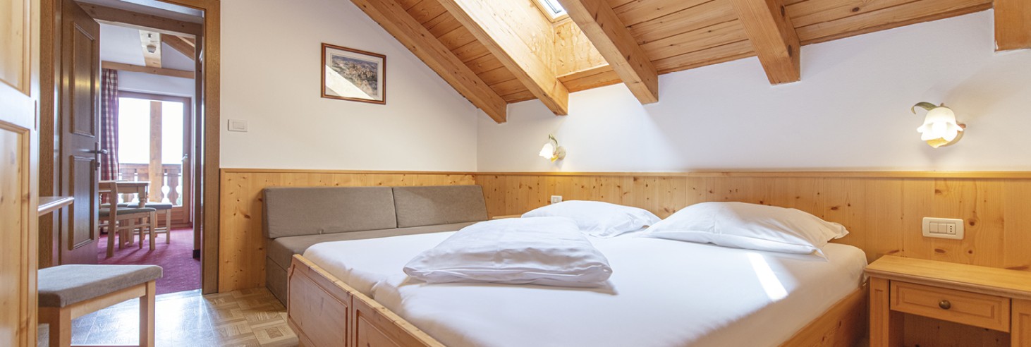 Rifugio Molignon Alm di Siusi | Le nostre camere vi invitano a rimanere La foto mostra la camera da letto con letto matrimoniale e divano. Dalla porta si vede la seconda camera da letto con balcone.