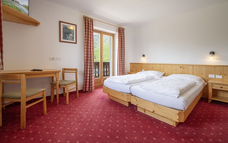 La foto mostra una camera matrimoniale con moquette, balcone, TV e scrivania.